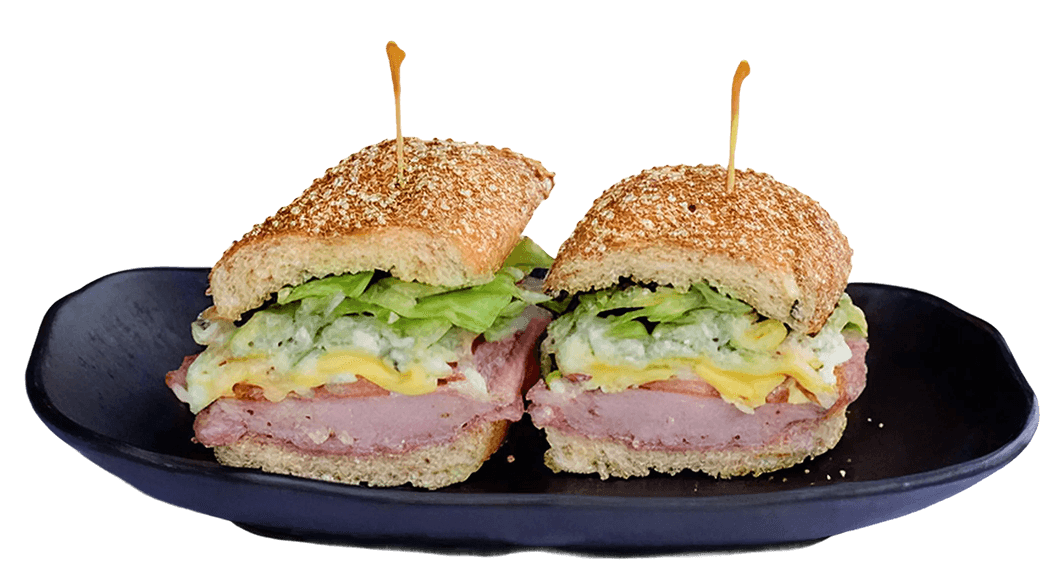 Multi-grain baguette sandwich filled with sliced chicken, yellow cheese, lettuce, pesto, and sun-dried tomatoes, served on a white plate.