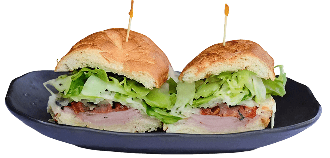 A hearty seeded sandwich filled with sliced chicken, crisp lettuce, cheese, pesto, and sundried tomatoes, served on a white plate.