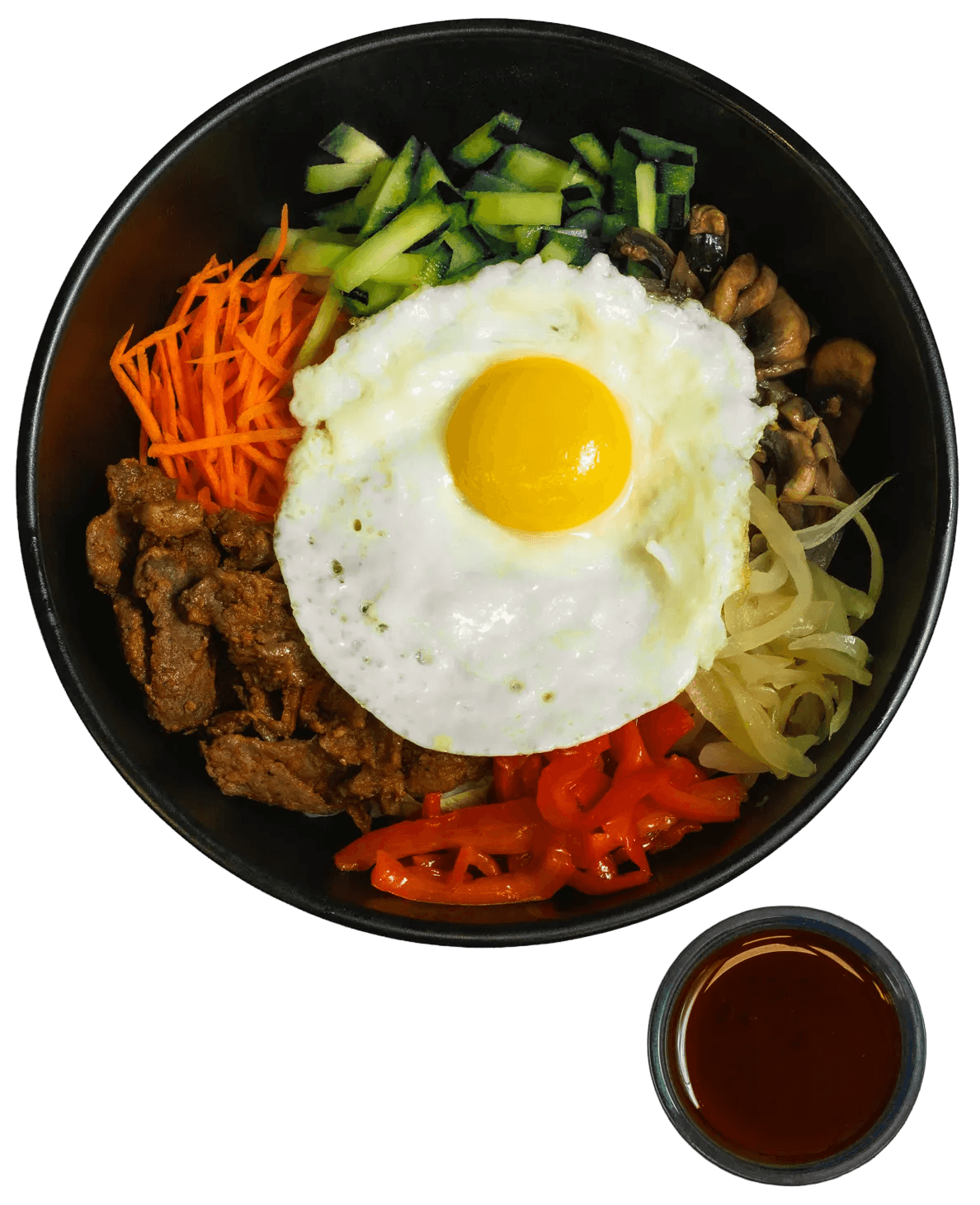 A top-down view of a black bowl of bibimbap, featuring a sunny-side-up egg, strips of beef, julienned carrots, chopped zucchini, sautéed mushrooms, sliced onions, and red peppers, with a small cup of sauce beside it.